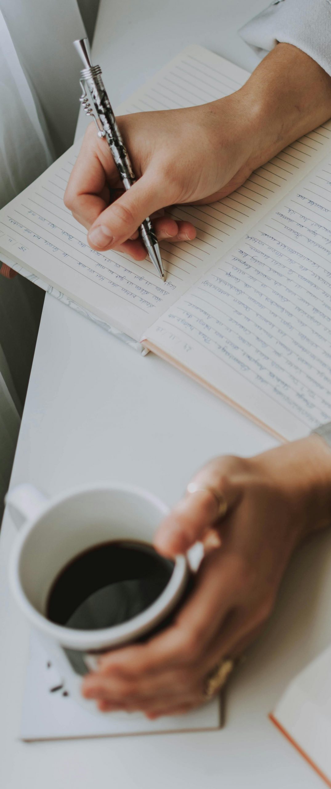 Close-up of a person writing in a notebook with coffee on a desk. Ideal for workplace or study themes.